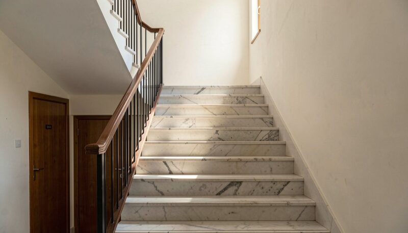 A marble entrance hall staircase with a wooden handrail, a shaft of daylight across the steps, an unmarked wooden door below